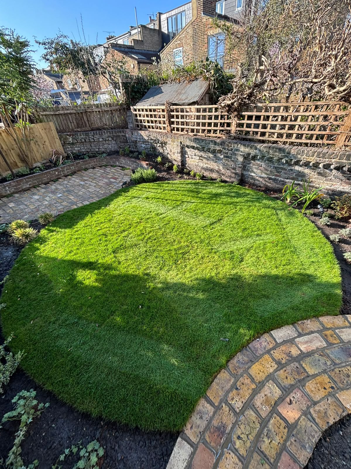 A lush oval lawn in sunlight, edged by a weathered brick path and planted borders.