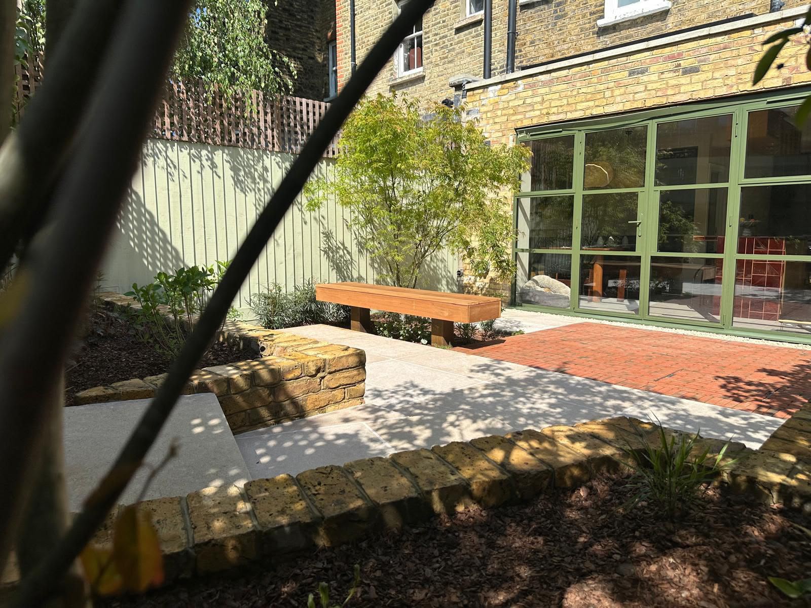 A quiet courtyard with a timber bench, pale stone paving, and sage-green framed glass doors.